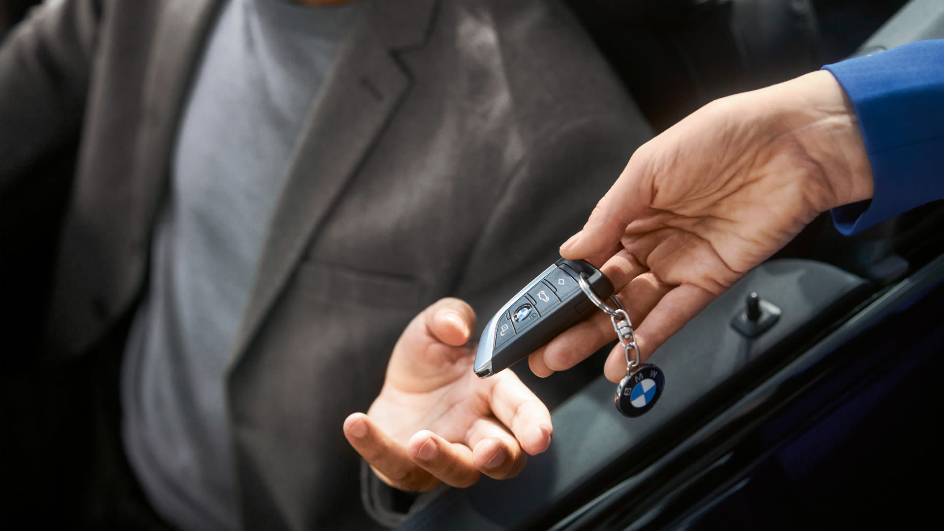 Person in gray blazer receiving a BMW key fob from another person in blue sleeve, inside a car.