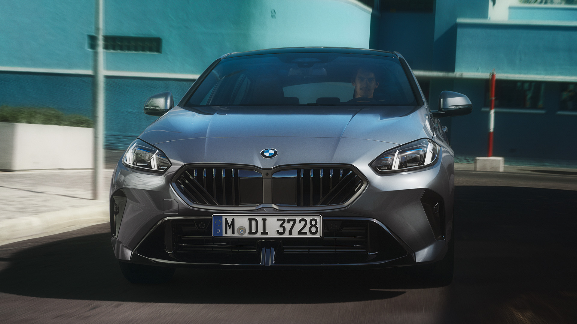 Front view of a silver BMW car with license plate M DI 3728 driving on a street with modern blue buildings and mountains in the background.