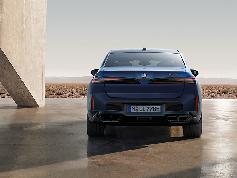 Rear view of a blue BMW 7 Series Saloon parked on a paved surface with a desert landscape in the background.