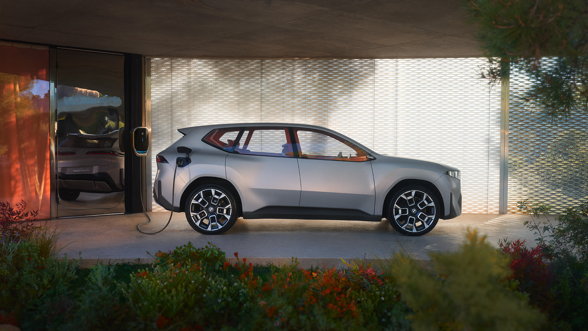 BMW electric SUV charging at a modern home station with glass architecture, metal mesh design, and landscaped garden