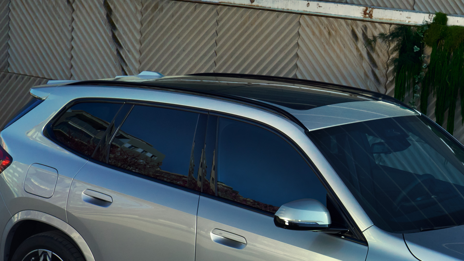 Close-up view of the upper part of a silver SUV with tinted windows and a panoramic sunroof, parked in front of a corrugated metal wall with some greenery.