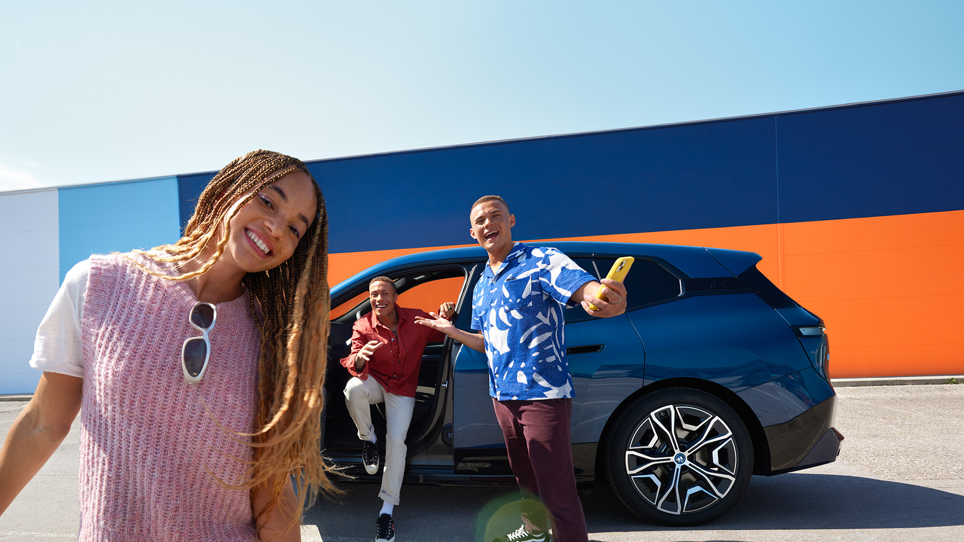 Three people standing near a blue car with one person sitting inside, in front of a colorful wall.