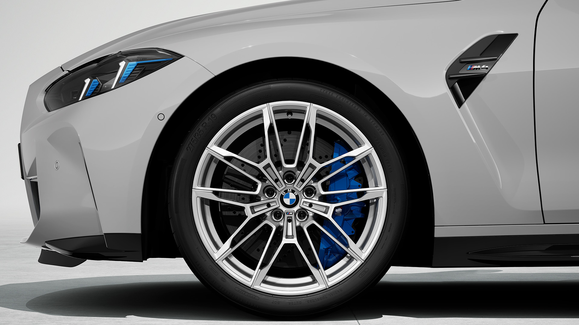 Close-up view of the front left wheel and part of the front end of a gray BMW car, showcasing the alloy rim, blue brake caliper, and M badge on the side vent.