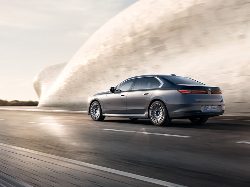 BMW 7 Series Saloon in metallic grey driving past a curved modern stone building on a wet road.