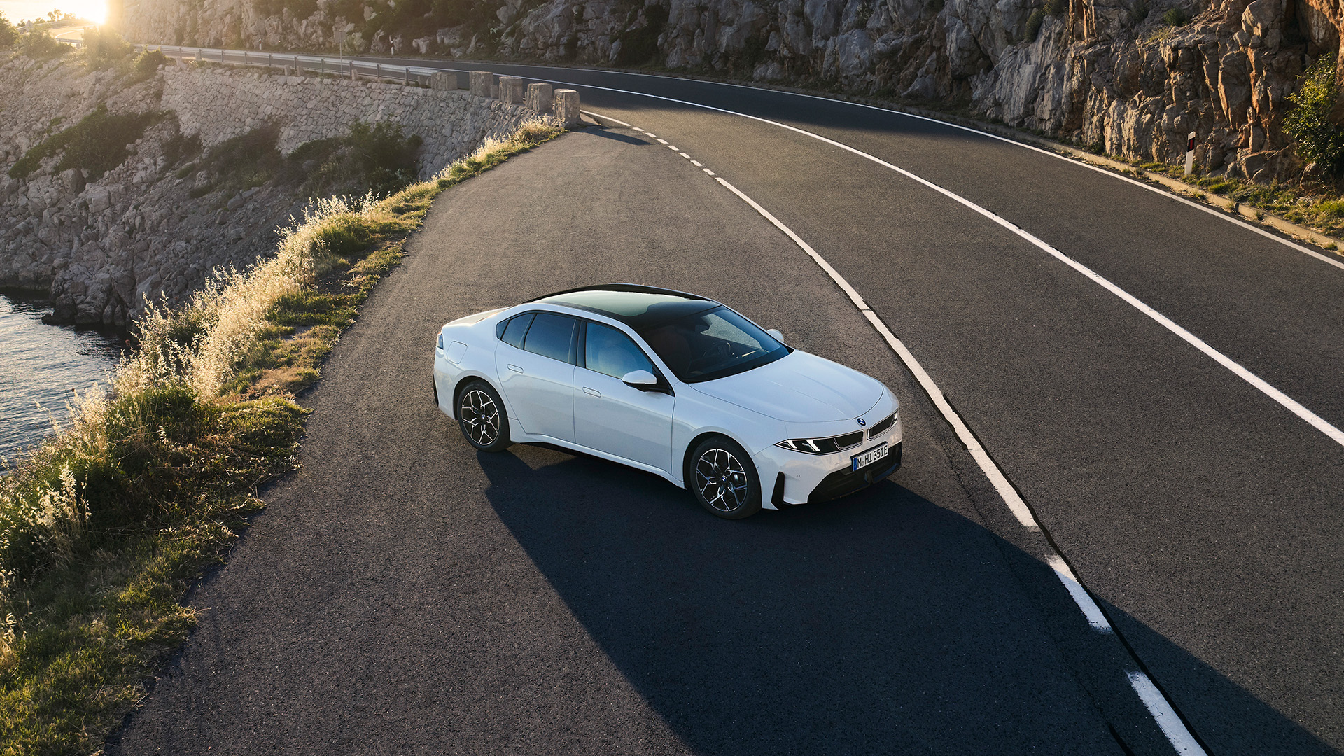 White BMW sedan parked on a winding coastal road at sunset, with rocky cliffs and sea views nearby.