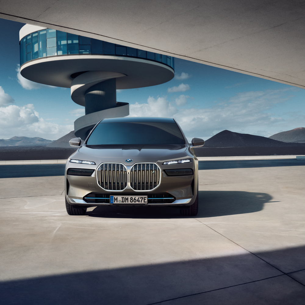 Front view of a BMW car with a modern spiral-designed building and large windows in the background, set against a mountainous landscape under a blue sky.