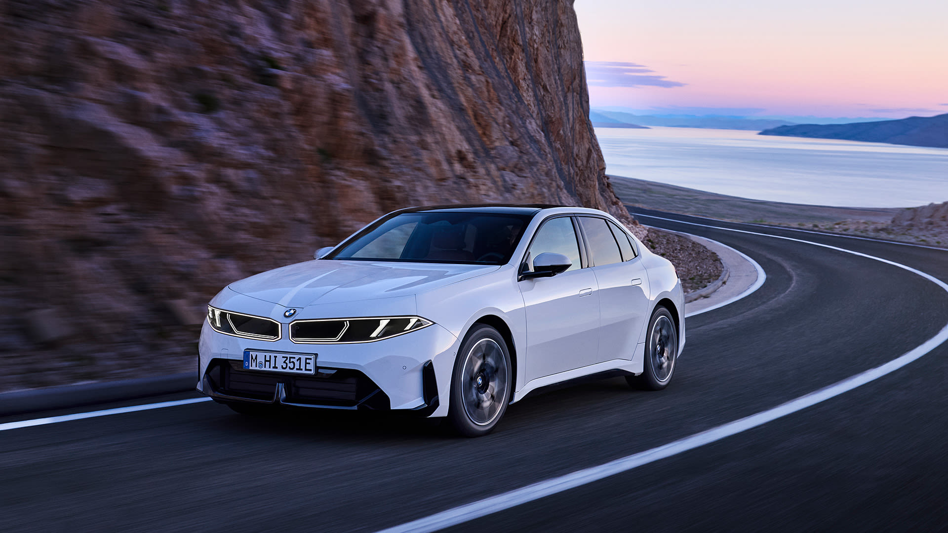 White BMW i3 sedan drives along a winding coastal mountain road at dusk, with cliffs and water in the background.