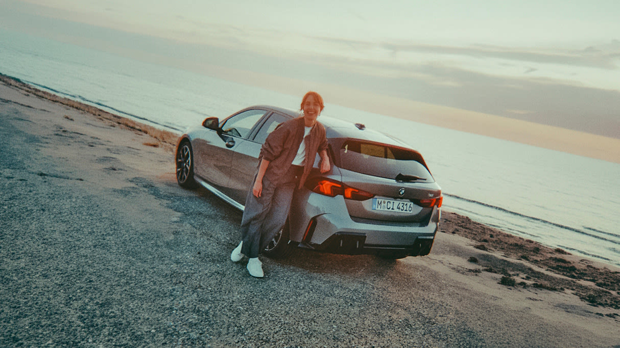 Person leaning against a silver BMW 1 Series hatchback parked on a coastal road, with the sea and sunset horizon in the background.