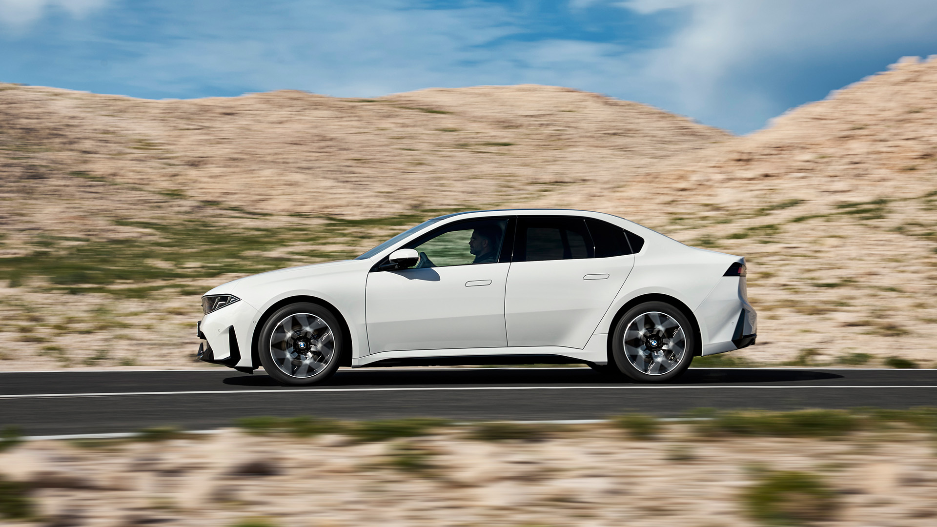 White BMW sedan driving on a desert road, with motion blur and rocky hills in the background.
