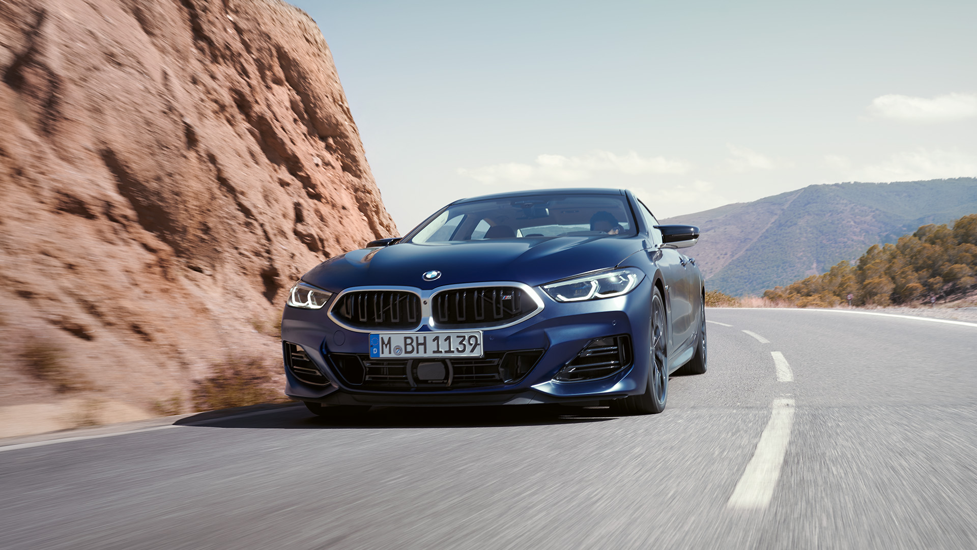 A sleek, dark-colored BMW car driving on a scenic mountain road with rocky cliffs on the left and hills in the background under a clear sky.