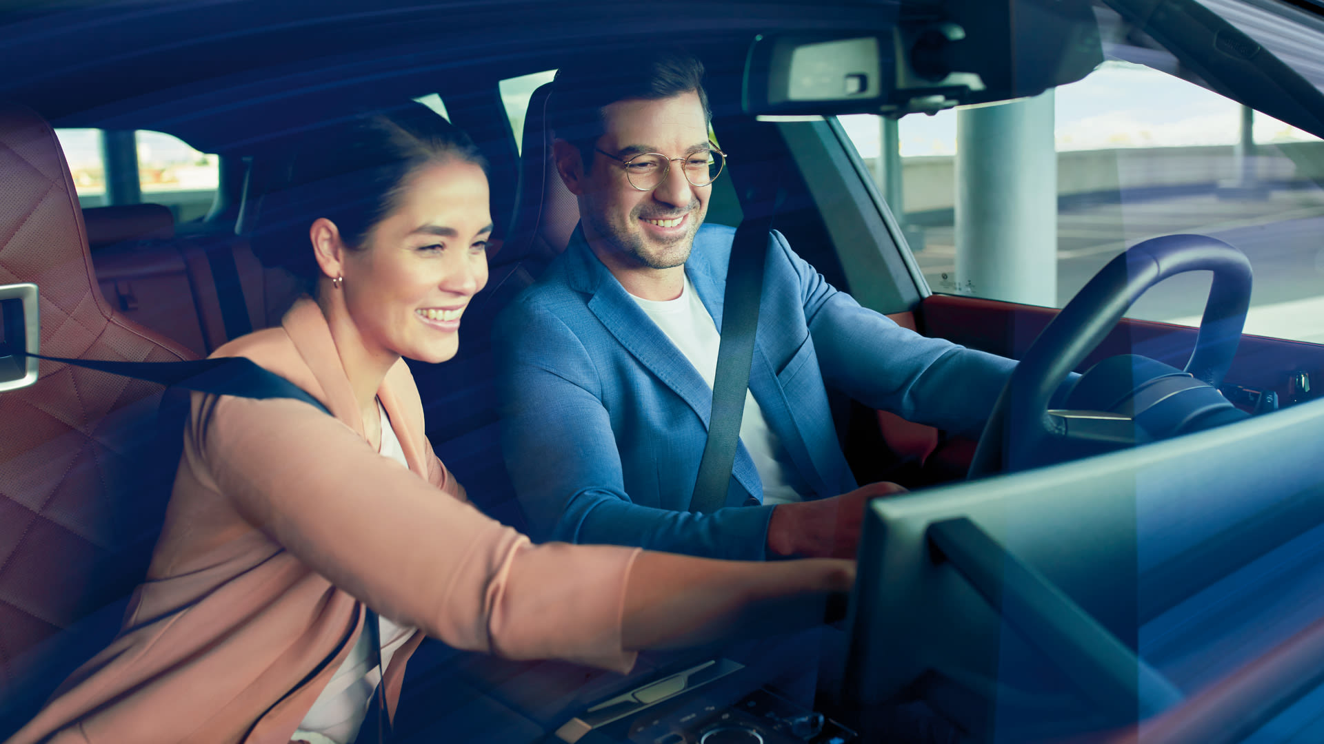 Two people sitting in the front seats of a car, both wearing seat belts; the person on the left is reaching toward the center console.