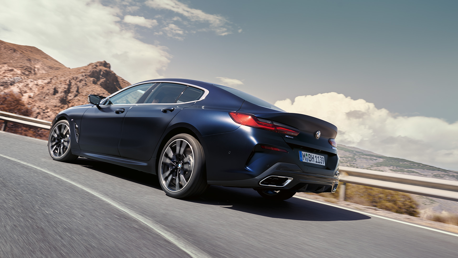 A sleek, dark-colored luxury BMW car driving on a winding mountain road with a scenic background of rocky hills and a partly cloudy sky.