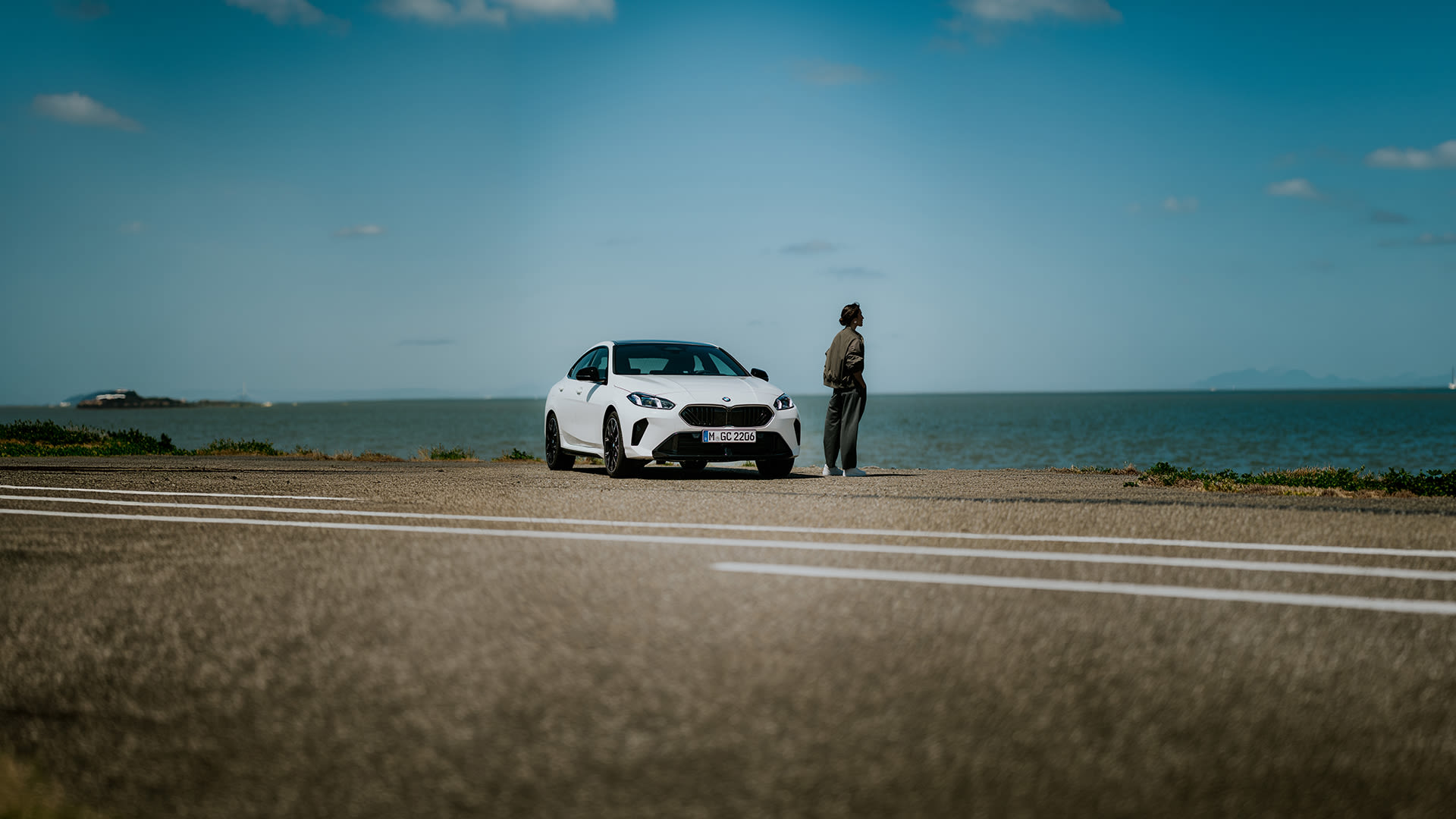 Person standing next to a white BMW 2 Gran Coupe parked by the seaside at sunset.