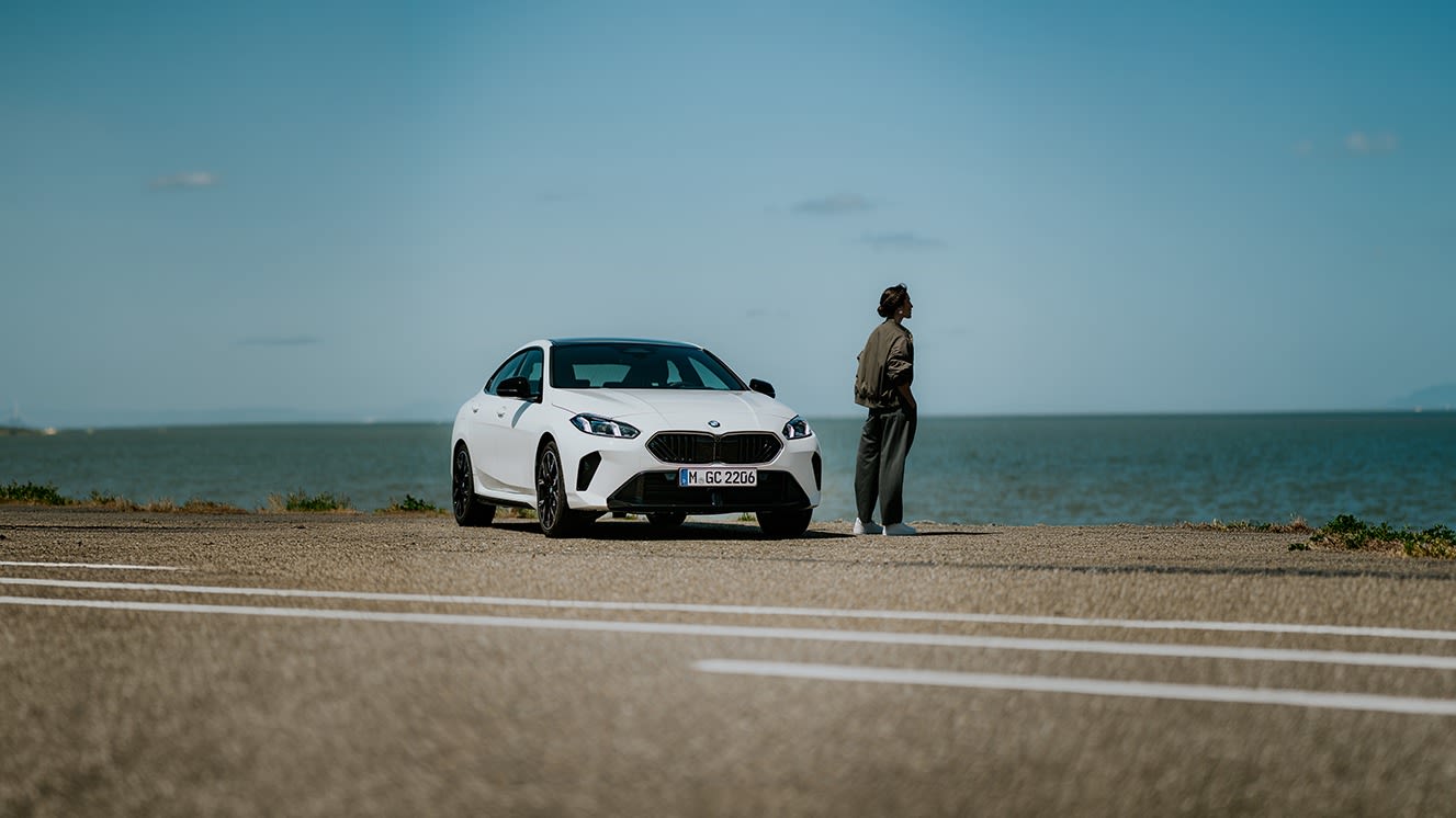 Person standing next to a white BMW 2 Gran Coupe parked by the seaside at sunset.