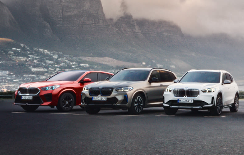 Three BMW SUVs parked side by side on a road with a mountainous landscape in the background. The cars are red, gray, and white.