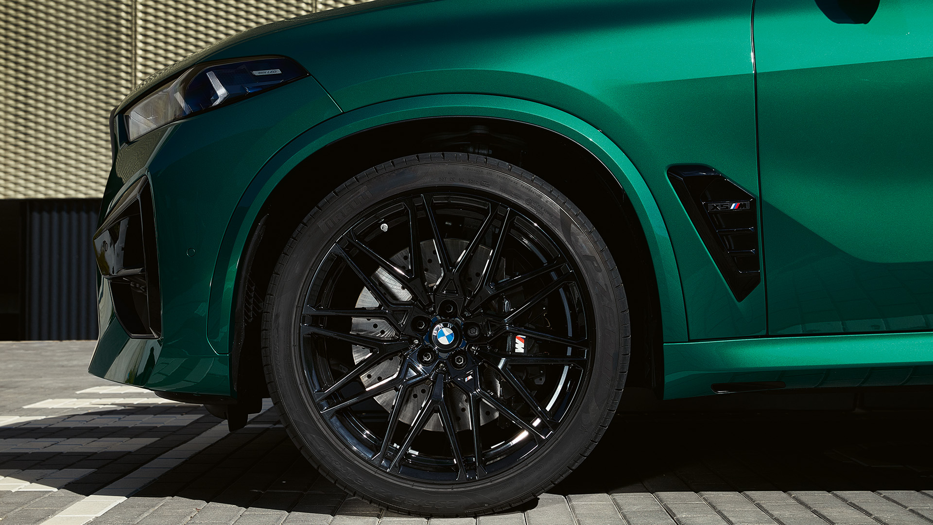 Close-up of the front side of a green BMW car with black alloy wheels, parked on a paved surface in front of a textured wall.