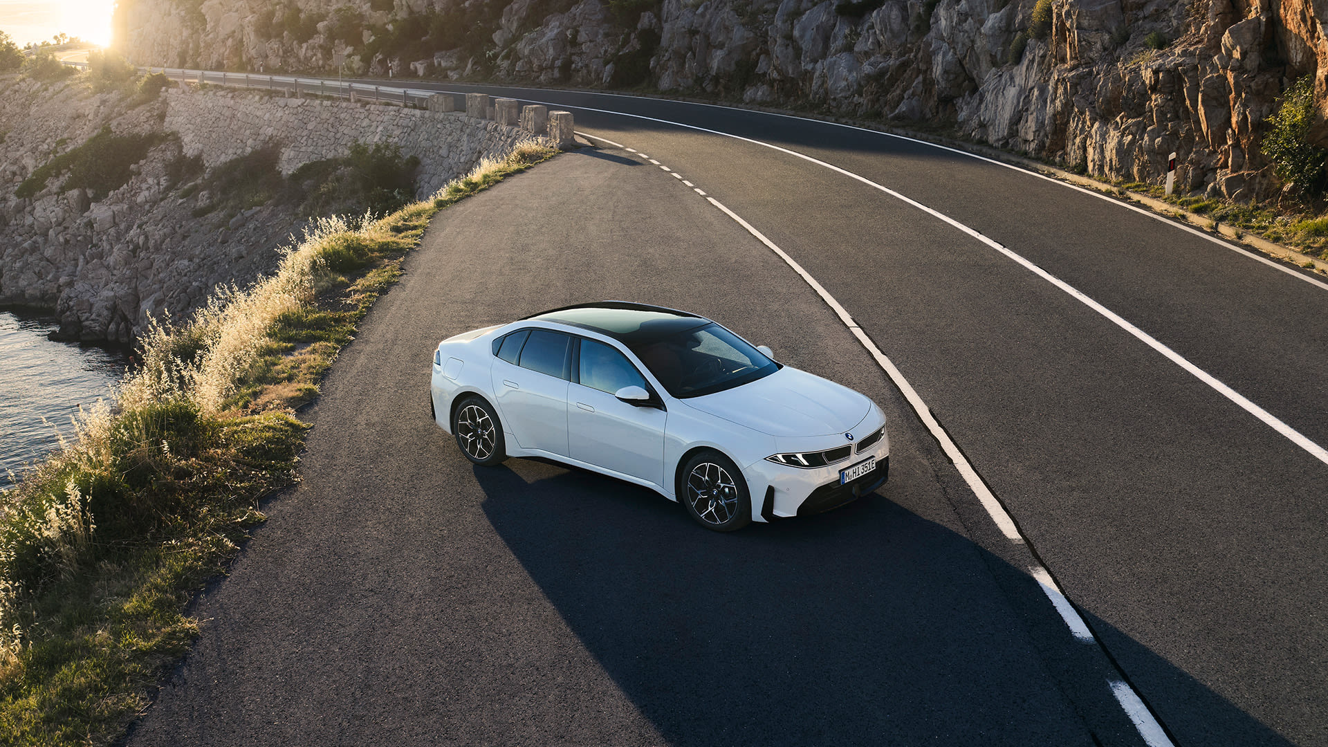 White all-electric BMW i3 Saloon parked on a coastal road at sunset, overlooking rocky cliffs and the sea.