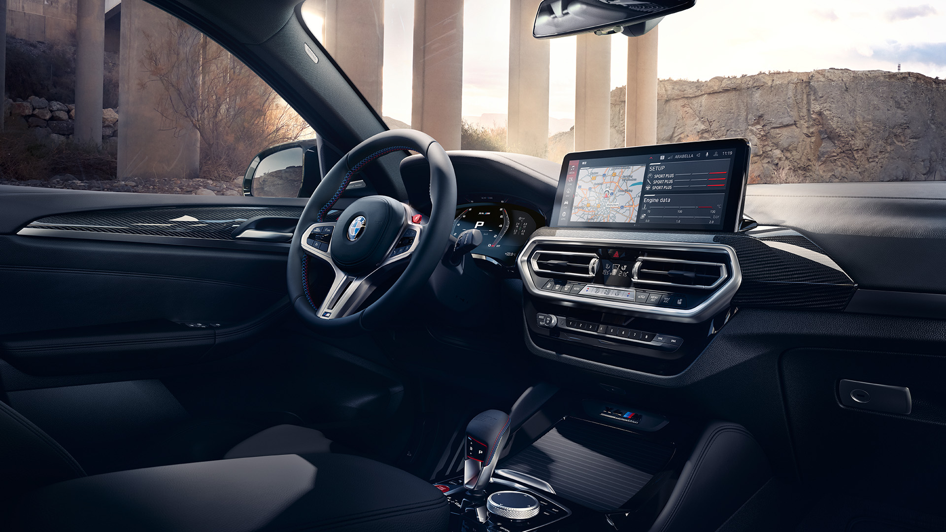 Interior of a modern car showing the steering wheel, dashboard, and infotainment system with a map displayed on the screen. The background outside the car window shows large concrete pillars and a rocky landscape.