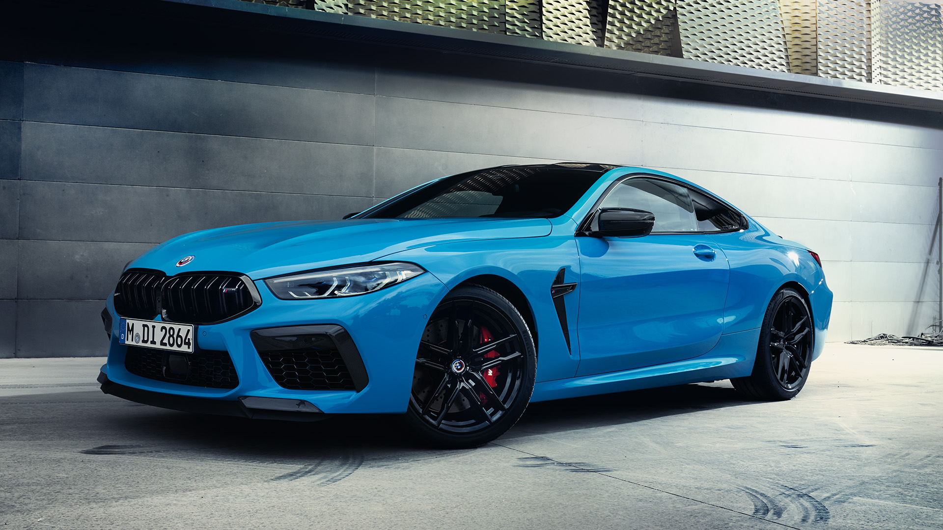 Blue BMW M8 Coupé car parked in an industrial setting with a modern metallic wall and geometric patterns in the background.