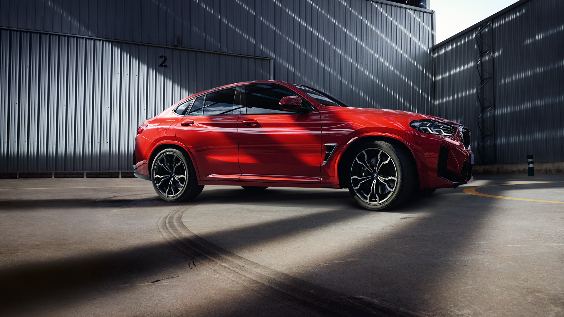Red BMW car parked in an industrial area with metal buildings and sunlight casting shadows.
