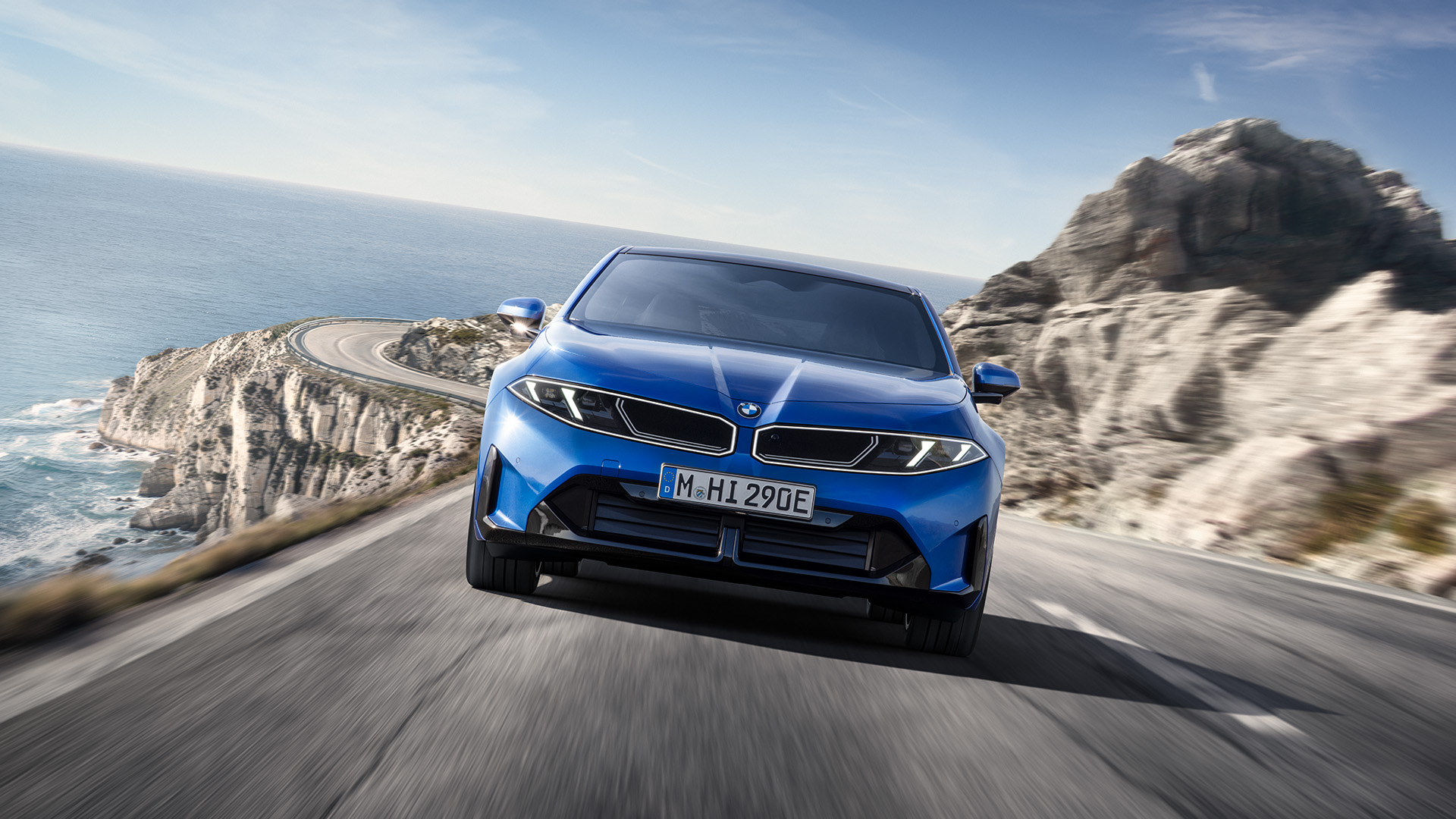 Blue BMW electric sedan driving along a winding coastal road, with ocean views and rocky cliffs in the background.