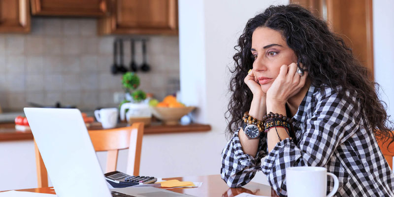 A woman sits at her kitchen table, looking forlornly at her computer.