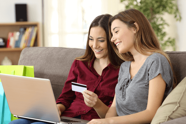 Two young women holding a credit card and looking at their laptops. 