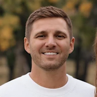 A smiling young man with short brown hair wearing a white sweater, photographed outdoors with blurred autumn foliage background.