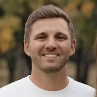 A smiling young man with short brown hair wearing a white sweater, photographed outdoors with blurred autumn foliage background.