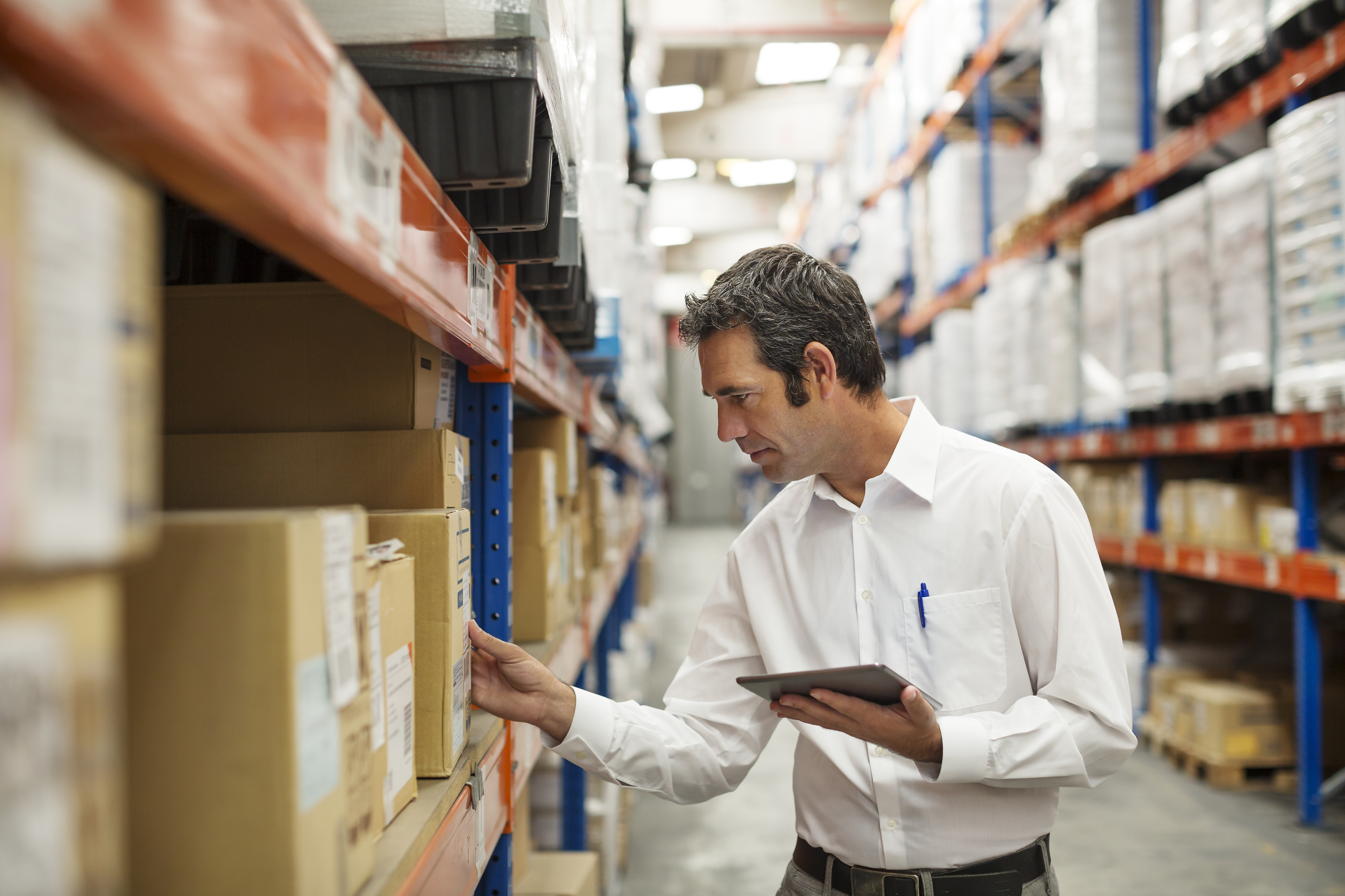Warehouse manager in a white shirt reviewing inventory with a tablet beside shelves of boxes