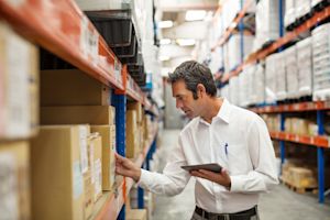 Warehouse manager in a white shirt reviewing inventory with a tablet beside shelves of boxes
