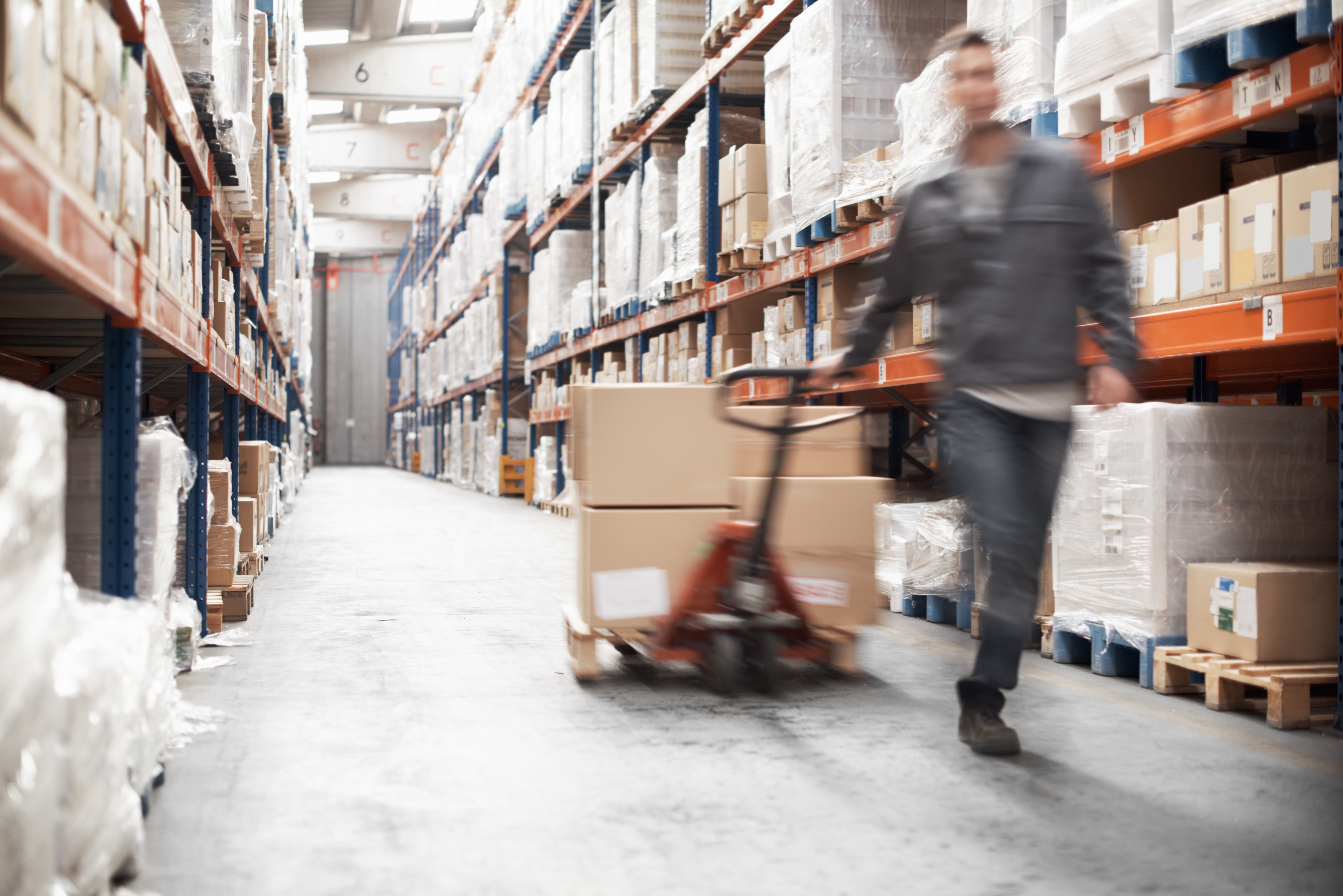 Warehouse associate pulling a pallet jack of boxes down an aisle of tall racks