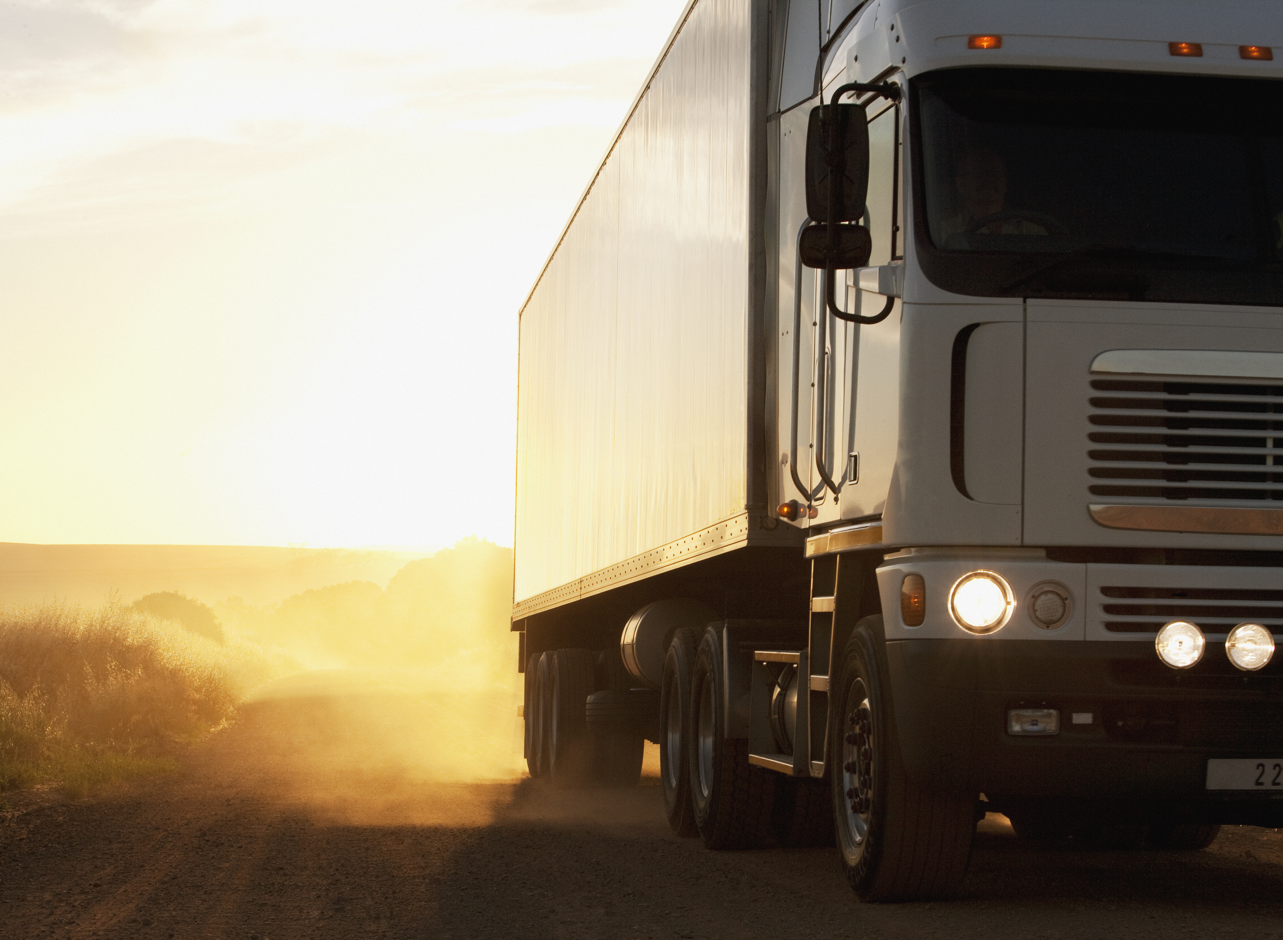 White semi-truck with a box trailer driving down a dusty rural road into a golden sunset