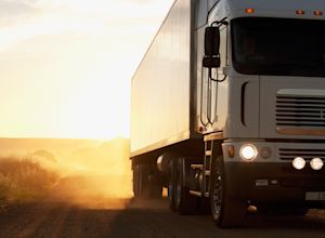 White semi-truck with a box trailer driving down a dusty rural road into a golden sunset