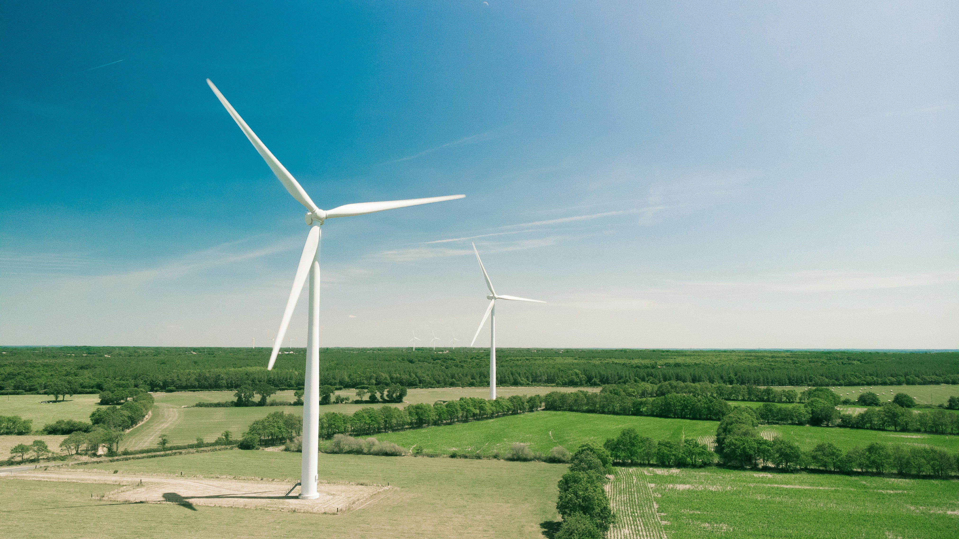 Two wind turbines on a green farmland landscape under a blue sky with distant turbines on the horizon