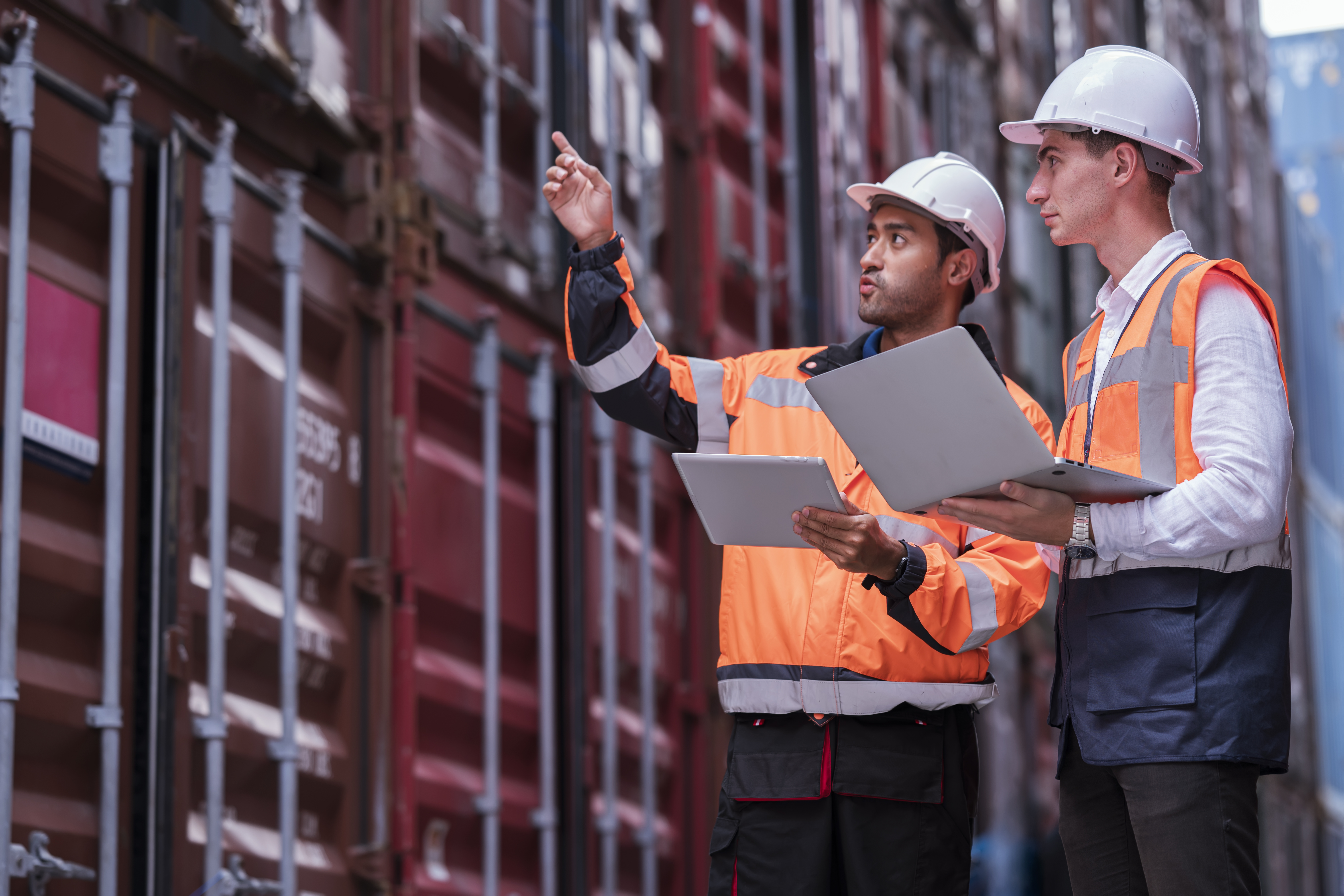 Two customs and logistics workers in safety vests and hard hats inspecting shipping containers at a port with a laptop