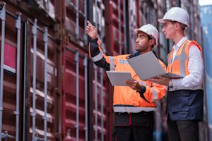 Two customs and logistics workers in safety vests and hard hats inspecting shipping containers at a port with a laptop