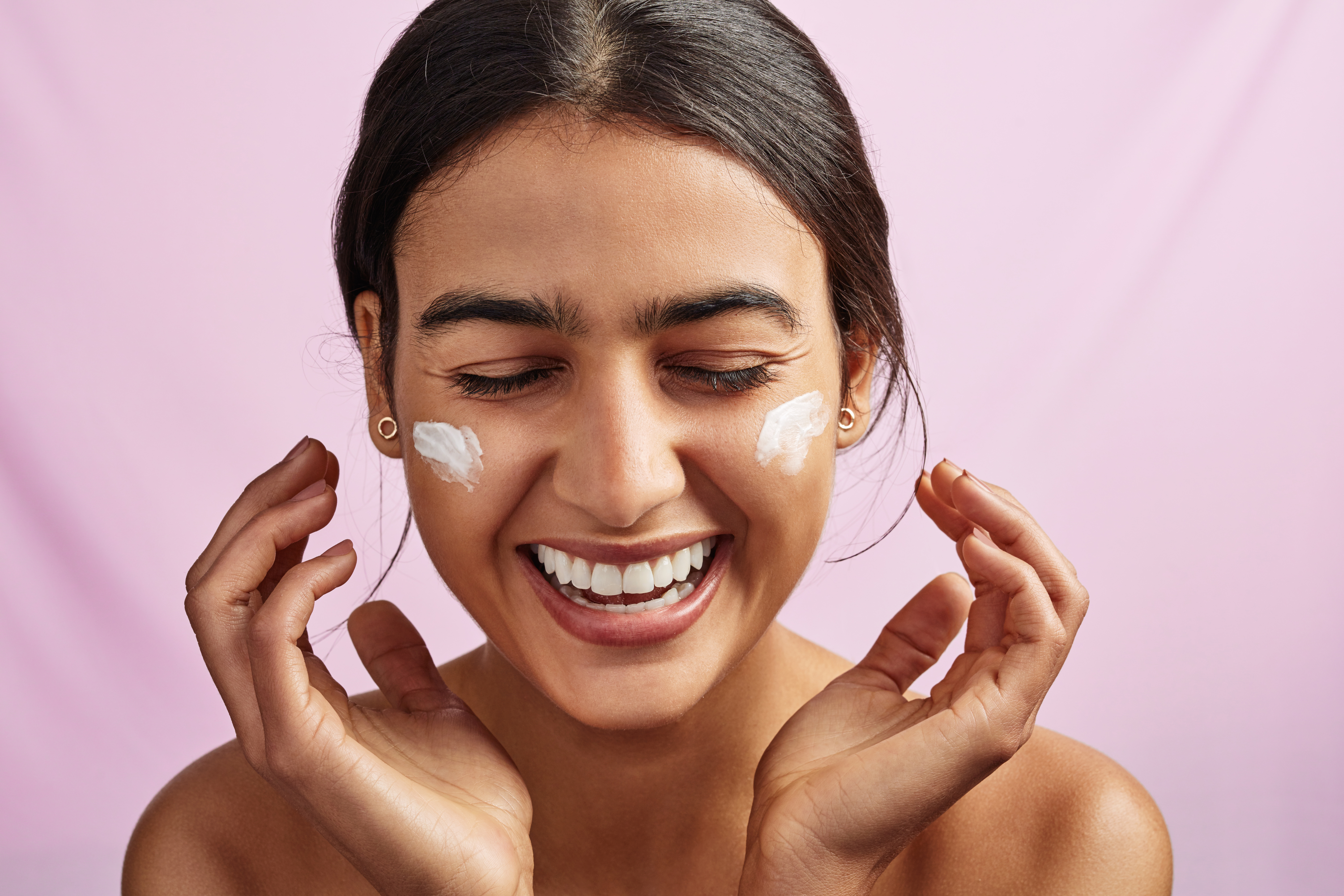 Smiling woman applying skincare cream to her face on a pink background