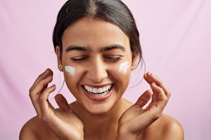 Smiling woman applying skincare cream to her face on a pink background