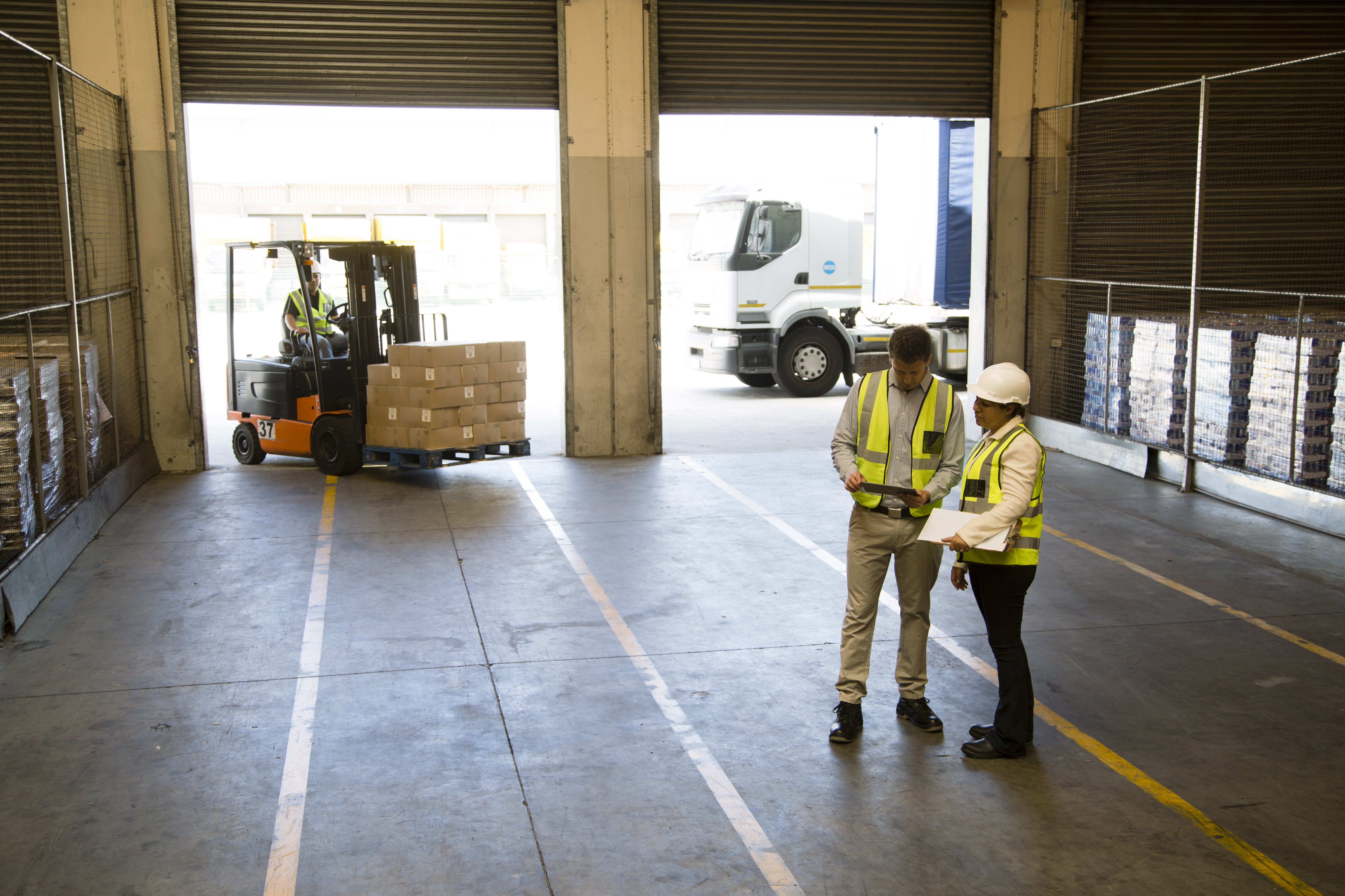 Two warehouse workers in hi-vis vests and hard hats talking on a loading dock as a forklift loads pallets onto a truck in the background
