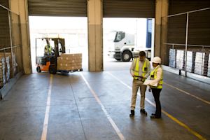 Two warehouse workers in hi-vis vests and hard hats talking on a loading dock as a forklift loads pallets onto a truck in the background