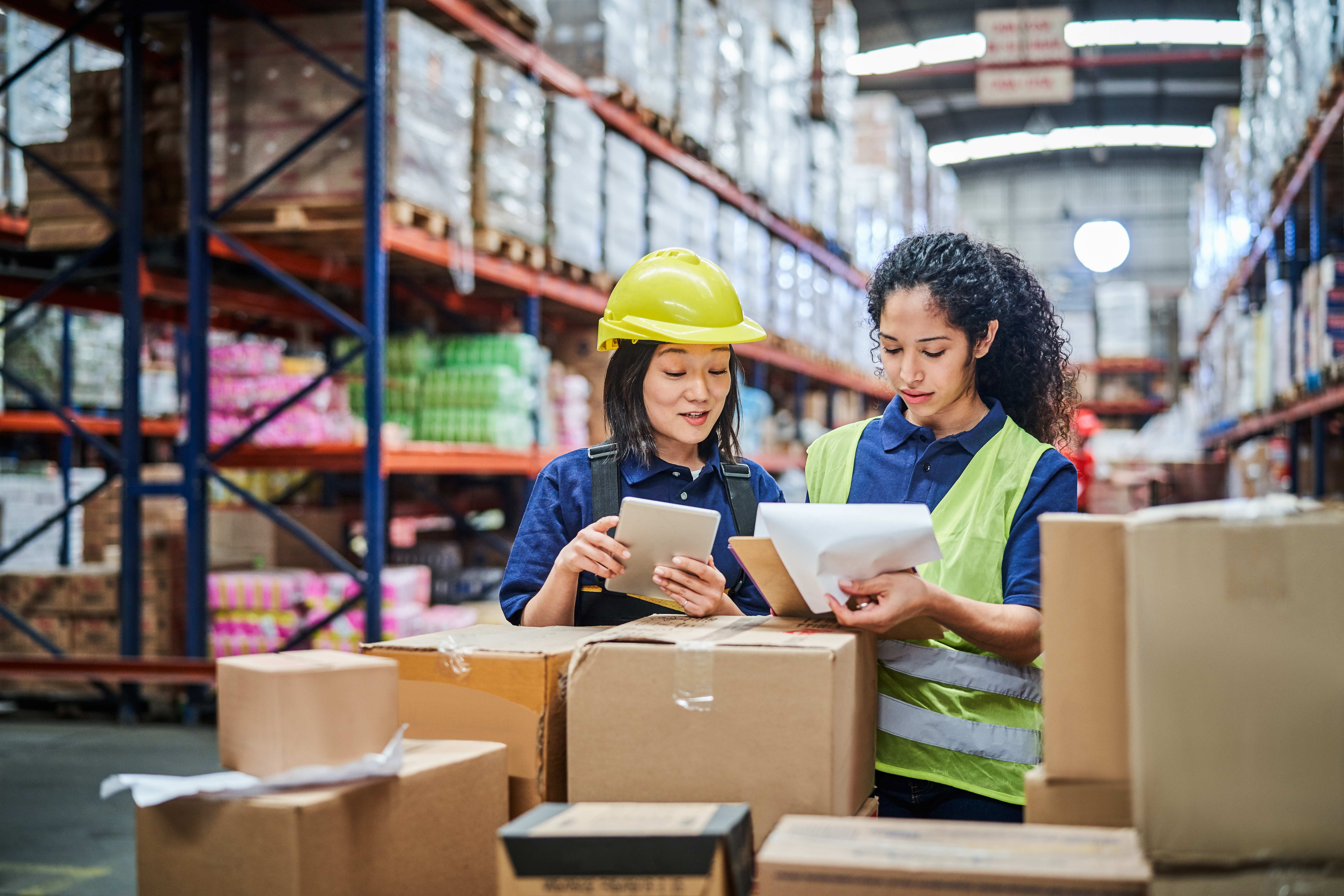 Two warehouse workers in hard hats and safety vests reviewing inventory on a tablet and clipboard amid cardboard boxes