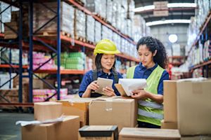 Two warehouse workers in hard hats and safety vests reviewing inventory on a tablet and clipboard amid cardboard boxes