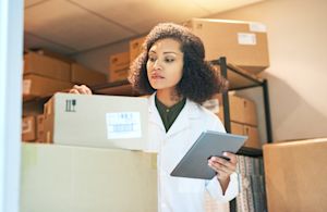 Pharmaceutical warehouse worker in a white lab coat inspecting a labeled shipping box while holding a tablet, surrounded by stacked inventory on shelves