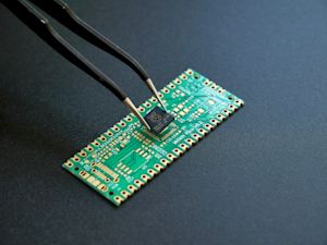 Tweezers placing a small semiconductor chip onto the solder pads of a green prototyping PCB