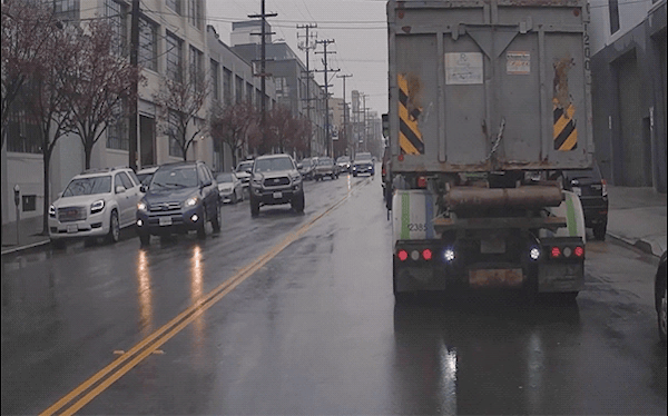 A Cruise AV navigates around a double-parked truck in the rain, with other vehicles approaching in the oncoming lane. The AV yields right-of-way to two vehicles, which in turn are going around a double-parked vehicle in their own lane.
