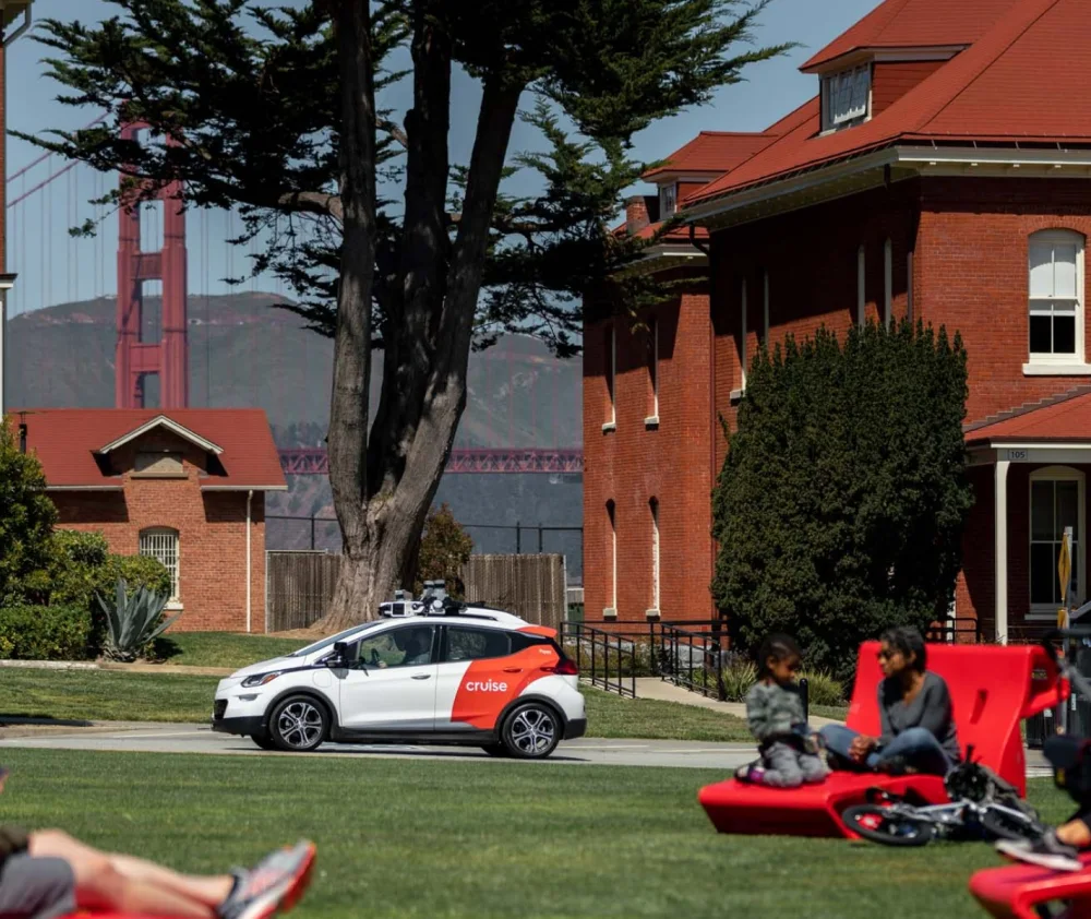 With a peek of the Golden Gate Bridge behind it, a Cruise first-generation AV drives past a park full of people enjoying a sunny day in San Francisco's Presidio.