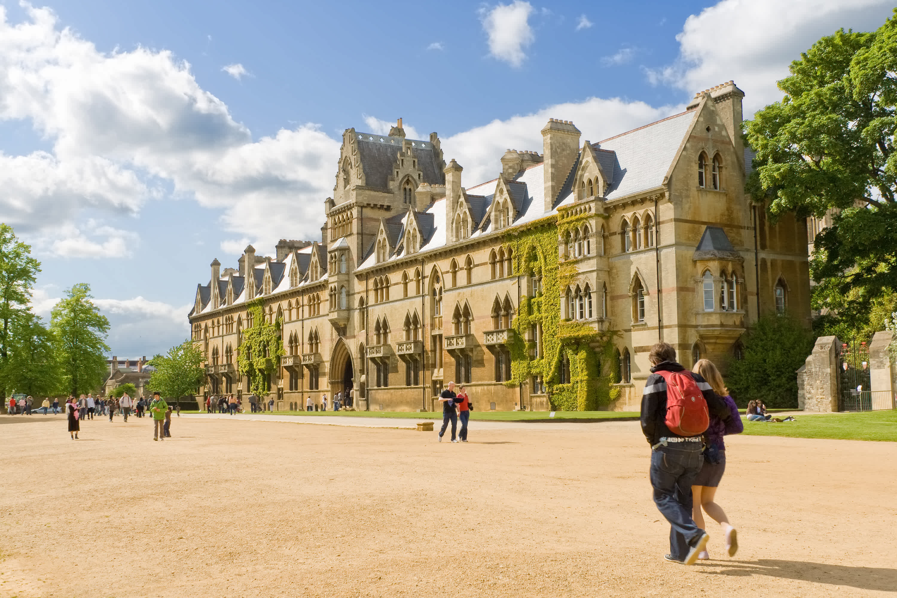 Several individuals stroll past a prominent university building, showcasing its architectural features against a bright sky. Several individuals stroll past a prominent university building, showcasing its architectural features against a bright sky.