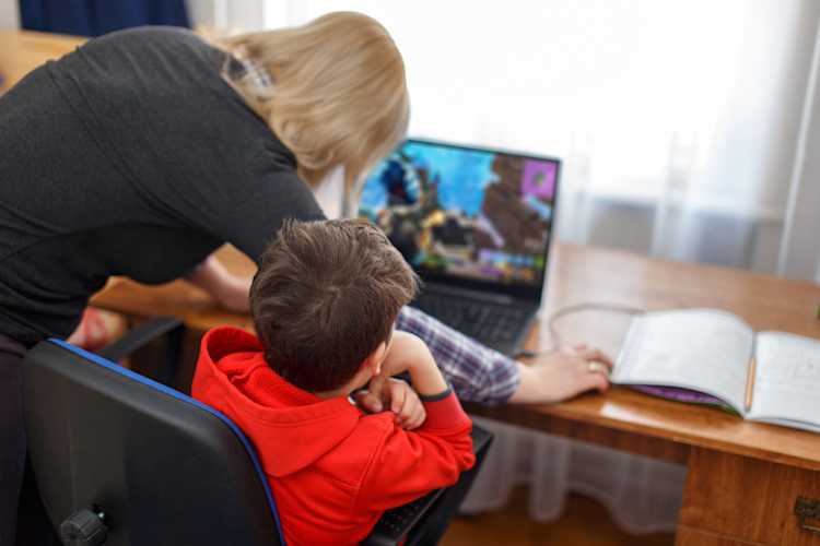 A woman standing behind a child sitting in front of a computer open to a gaming platform A woman standing behind a child sitting in front of a computer open to a gaming platform