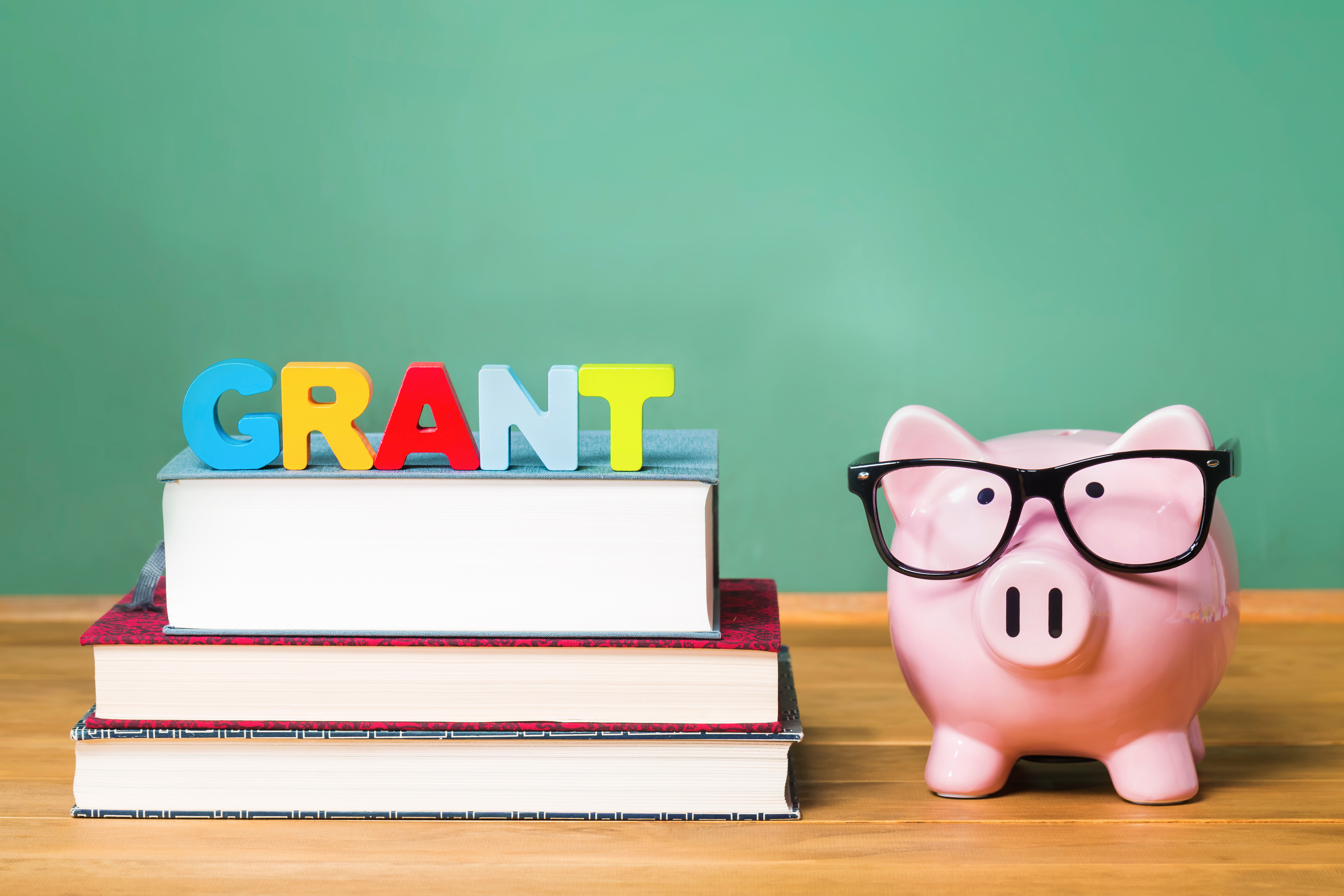 A wooden table displays a piggy bank and books, with a green board in the background, illustrating K-12 school safety grants.